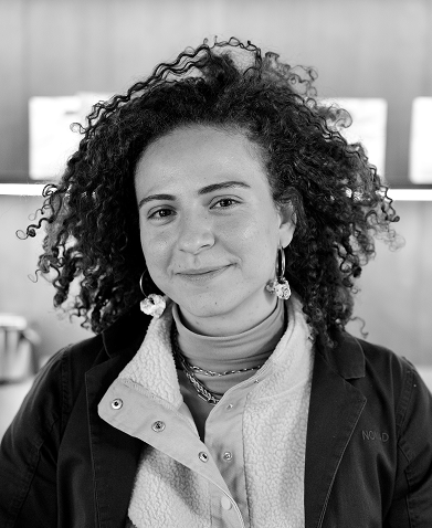 Retrato en blanco y negro de una persona con cabello rizado, vistiendo una chaqueta con el logotipo de NOMAD. La persona sonríe ligeramente, luce pendientes llamativos y varias capas de ropa, incluyendo una camiseta de cuello alto. El fondo muestra un entorno cálido, probablemente una cafetería.