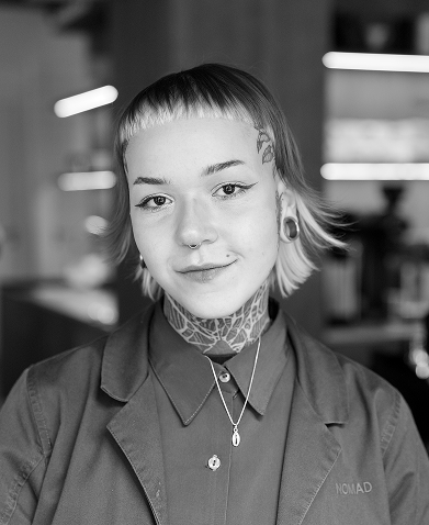 Retrato en blanco y negro de una persona con flequillo recto, cabello corto y teñido, y tatuajes visibles en el cuello y la cabeza. Lleva una chaqueta con el logotipo de NOMAD y una camisa abotonada. También lleva piercings en la nariz, orejas y labios, además de collares. La imagen está tomada en el interior de una cafetería, con un fondo desenfocado.